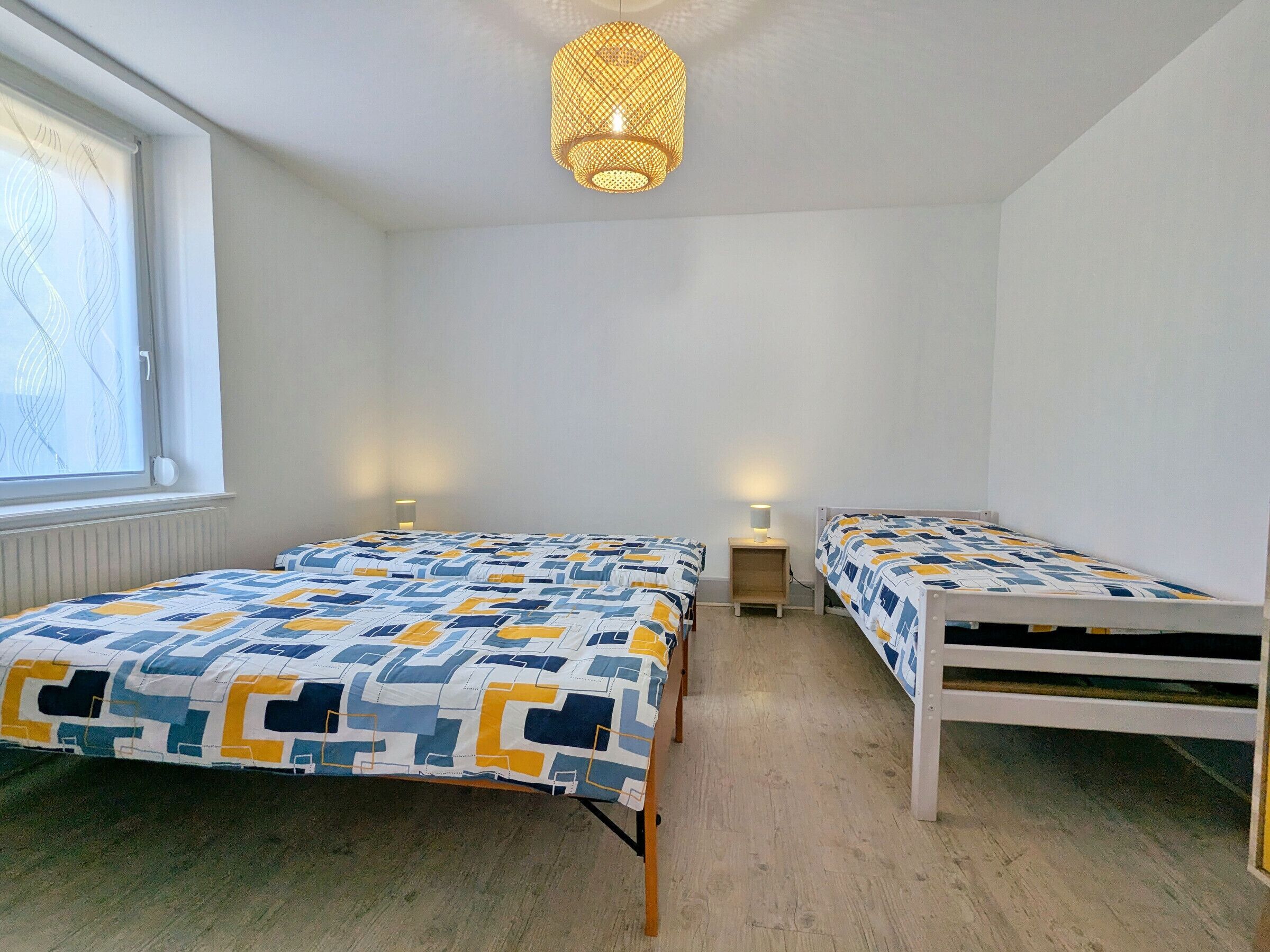 Et soveværelse med to senge, et vindue med persienner, en hængende lampe og et sengebord med en lampe.