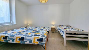 Et soveværelse med to senge, et vindue med persienner, en hængende lampe og et sengebord med en lampe.