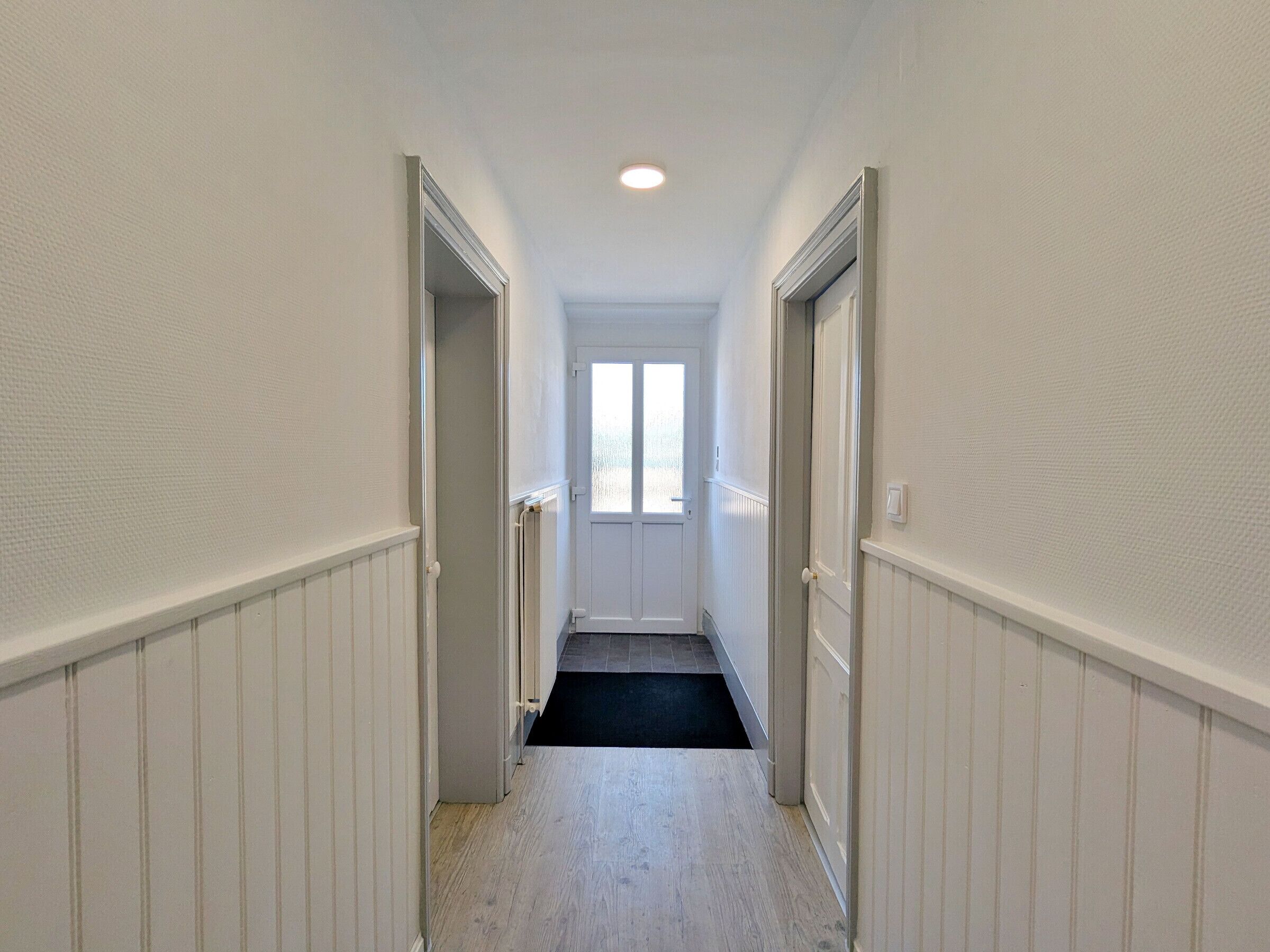 A narrow hallway with white walls and a black carpet leading to a door with a window.