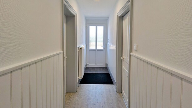 Un couloir étroit aux murs blancs et au tapis noir, qui mène à une porte dotée d’une fenêtre.