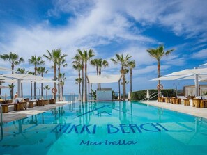 Ein klares, blaues Schwimmbecken, am Rand mit 'Marbella' beschriftet, umgeben von Palmen und Liegestühlen.