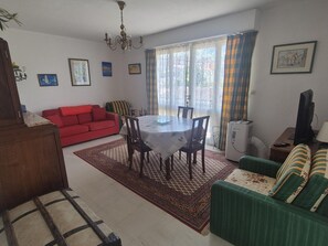 Ein Wohnzimmer mit einem roten Sofa, einem grünen Sofa, einem Esstisch mit Stühlen, einer Chandelierleuchtre und einem großen Fenster mit Vorhängen.