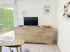 A room with a TV on a wooden cabinet, a small table, and a view of greenery outside. - T2 with private garden, calm - 4 guests (Saint-Georges-de-Didonne)