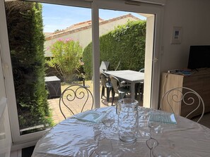 A room with a table set for dining, a grill outside, and a TV on a wooden cabinet. - T2 with private garden, calm - 4 guests (Saint-Georges-de-Didonne)