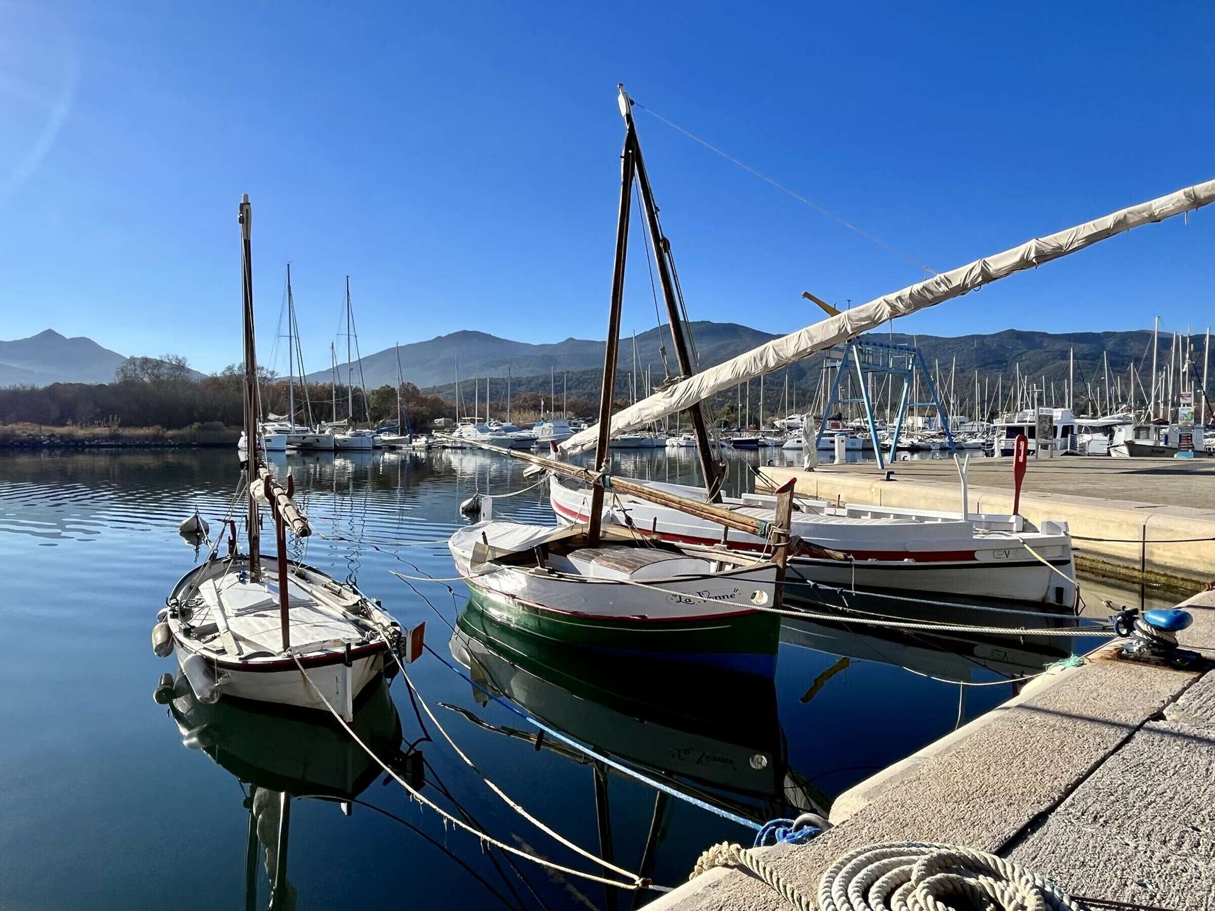 Un puerto deportivo con varias embarcaciones amarradas, agua tranquila y montañas al fondo.