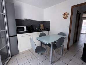 Een compacte keuken met witte kastjes, een magnetron, een broodrooster en een spoelbak. Er staat een glazen tafel met vier stoelen en een decoratieve klok aan de muur.