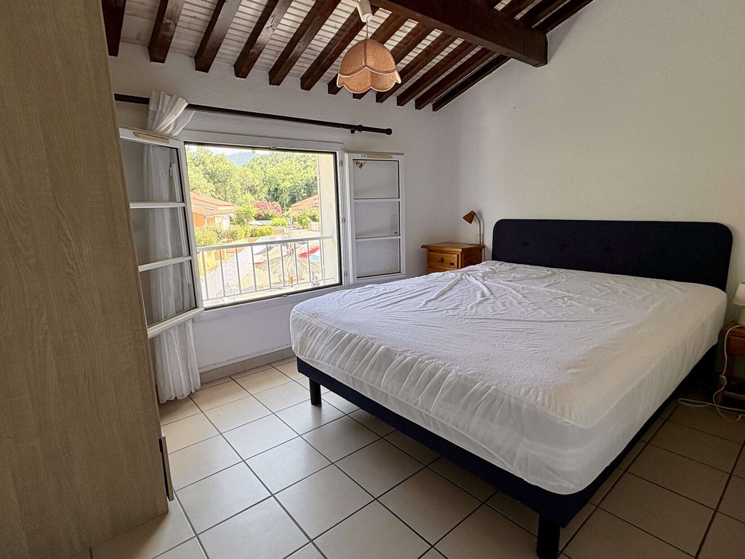 Un dormitorio con cama, ventana con cortinas, mesita de noche y ventilador de techo.