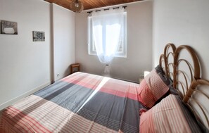 A bedroom with a bed, a window with a sheer curtain, a bedside table, and a wall-mounted lamp.