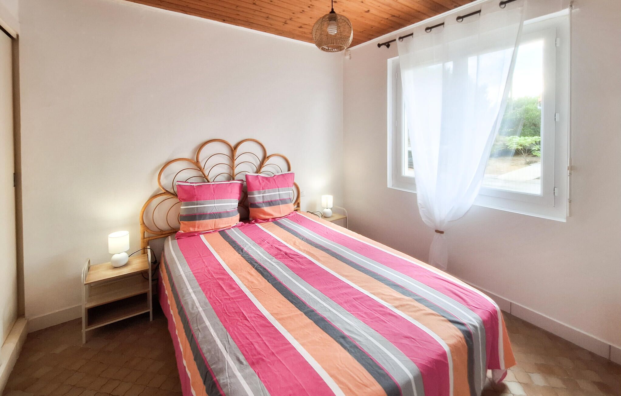 A bedroom with a bed, bedside lamps, a window with curtains, and a wooden ceiling.