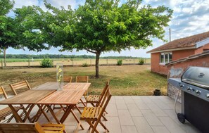 Un patio al aire libre con mesa y sillas de madera, una parrilla y un árbol grande que da sombra.