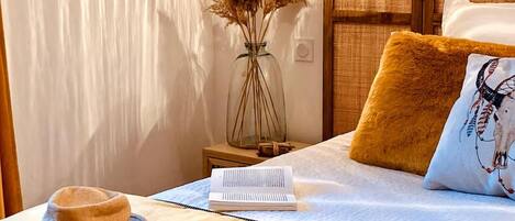 Ein ordentlich bezogenes Bett mit weißer Bettdecke, einem braunen Kopfkissen, einem Buch, einem Hut und einer Vase mit Trockenpflanzen auf einem Nachttisch.
