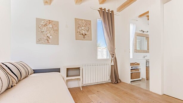 Un dormitorio con cama, un radiador, una ventana con cortinas y un baño visible a través de una puerta abierta.