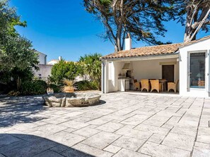 A covered outdoor patio area with stone flooring, a seating arrangement, and a view of the surrounding garden and trees. - Elegant house in nature - 6 guests (Saint-Clément-des-Baleines)