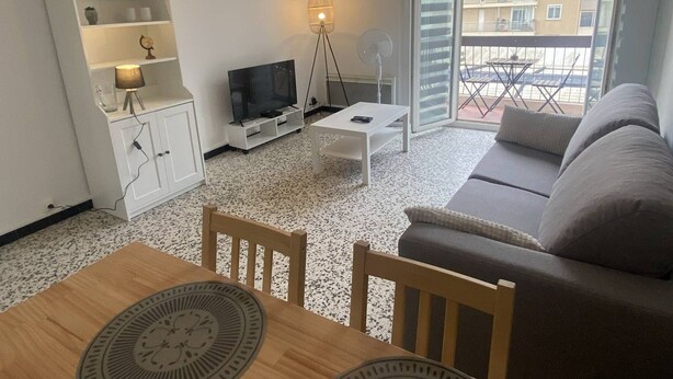 A living room with a TV, sofa, dining table, and a balcony with a view of buildings. - Cross apartment with parking - 4 guests (Canet-en-Roussillon)