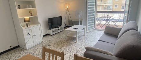 A living room with a sofa, a TV, a dining table with chairs, a cabinet, and a balcony with a view of buildings.