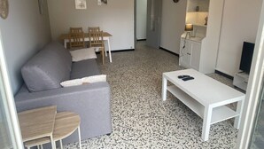 A compact living space with a grey sofa, a small wooden table, a white coffee table, a dining area with chairs, a kitchen area with a white cabinet, and a hanging light fixture. - Cross apartment with parking - 4 guests (Canet-en-Roussillon)
