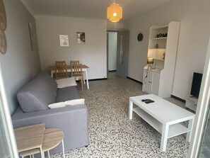 Ein kompakter Wohnbereich mit grauem Sofa, kleinem Holztisch, weißem Couchtisch, Essbereich mit Stühlen, Küchenzeile mit weißem Schrank und einer Pendelleuchte.