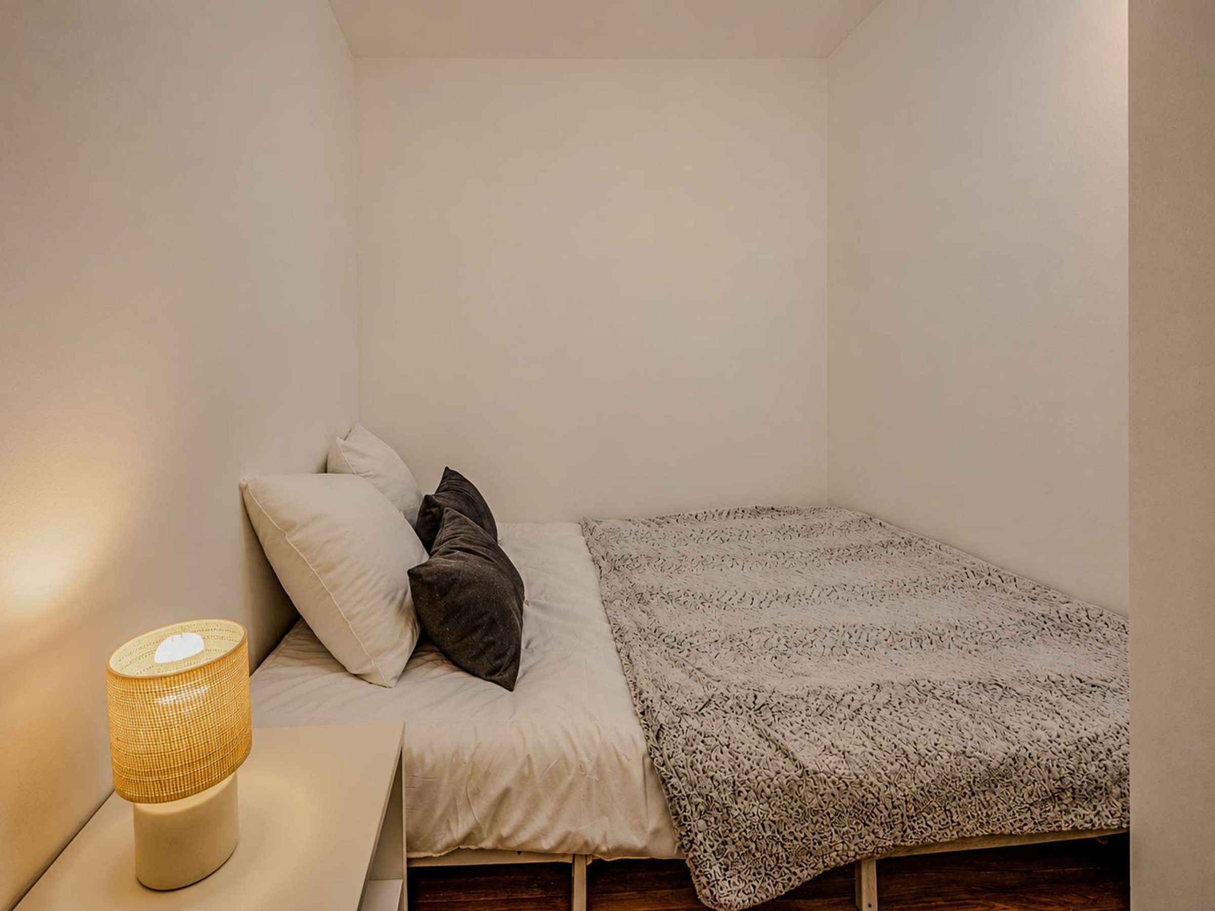 A small, single-bed room with a bedside table, a lamp, and a patterned blanket.