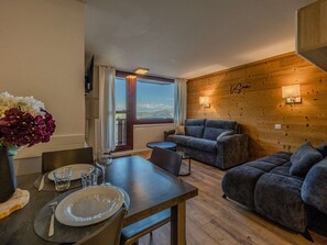 A cozy room with a wooden wall, a sofa, a dining table with plates and glasses, and a window with curtains. - Renovated apartment, ideal location - 6 guests (Plagne Bellecôte)