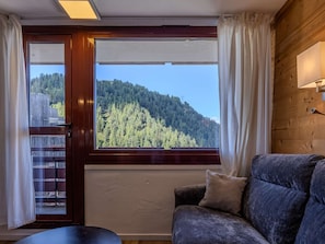 A room with a wooden interior, a sofa, a window with a view of a forested hill, and a door with a view of a fence. - Renovated apartment, ideal location - 6 guests (Plagne Bellecôte)