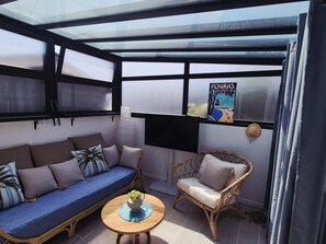 Une pièce lumineuse, avec un canapé, un fauteuil en osier, une petite table sur laquelle est posée une plante, et un téléviseur fixé au mur.
