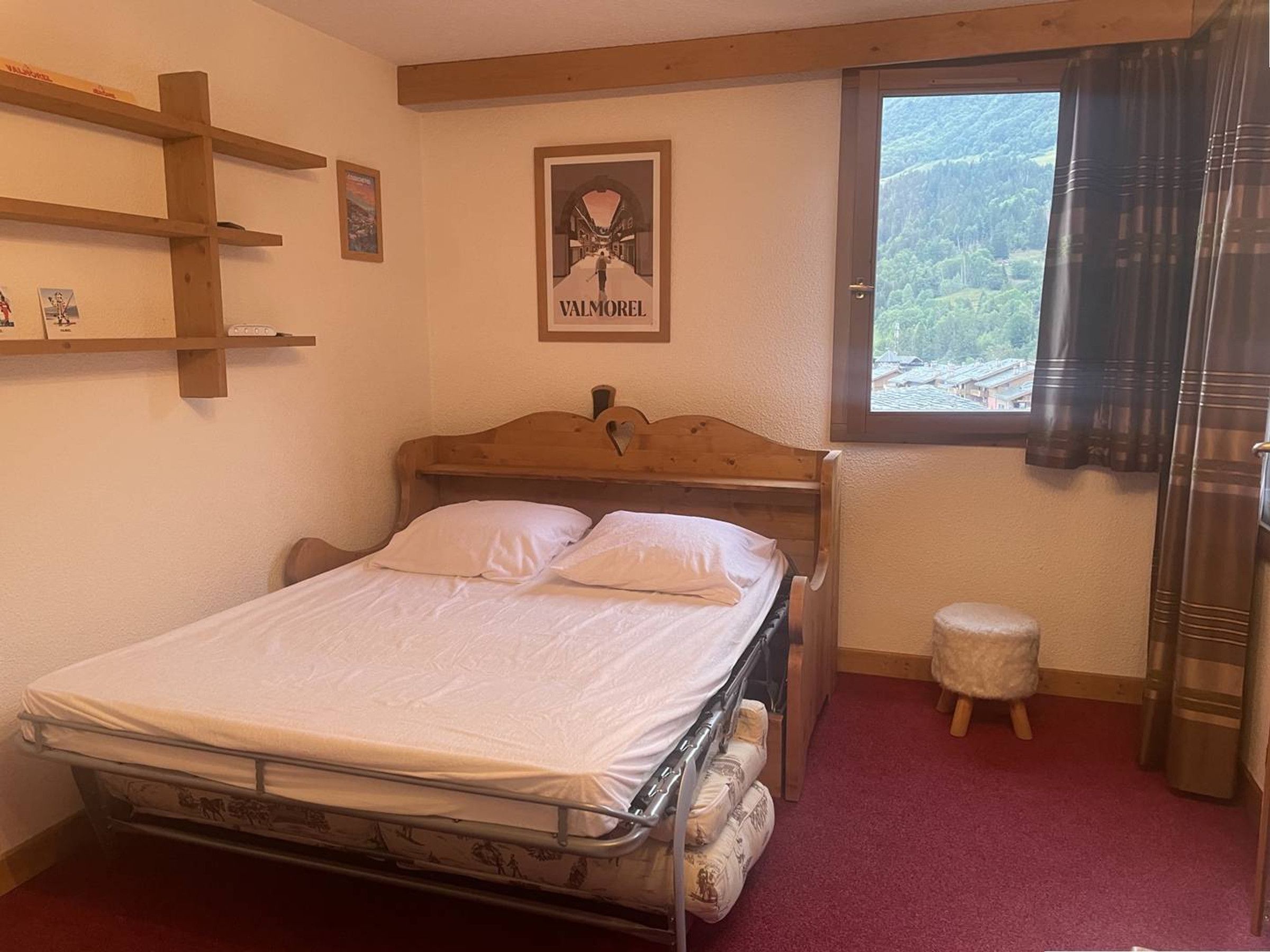 A bedroom with a wooden bed, a small stool, a window with a view of greenery, and a framed picture on the wall.