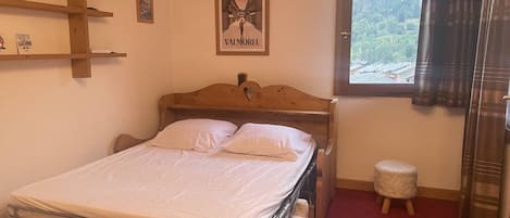 A bedroom with a wooden bed, a small stool, a window with a view of greenery, and a framed picture on the wall.