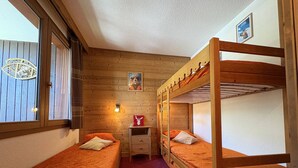 A room with two bunk beds, a small desk, a chair, a window with blinds, and a wall-mounted light fixture. - Apartment with station view - 2 guests (Valmorel)