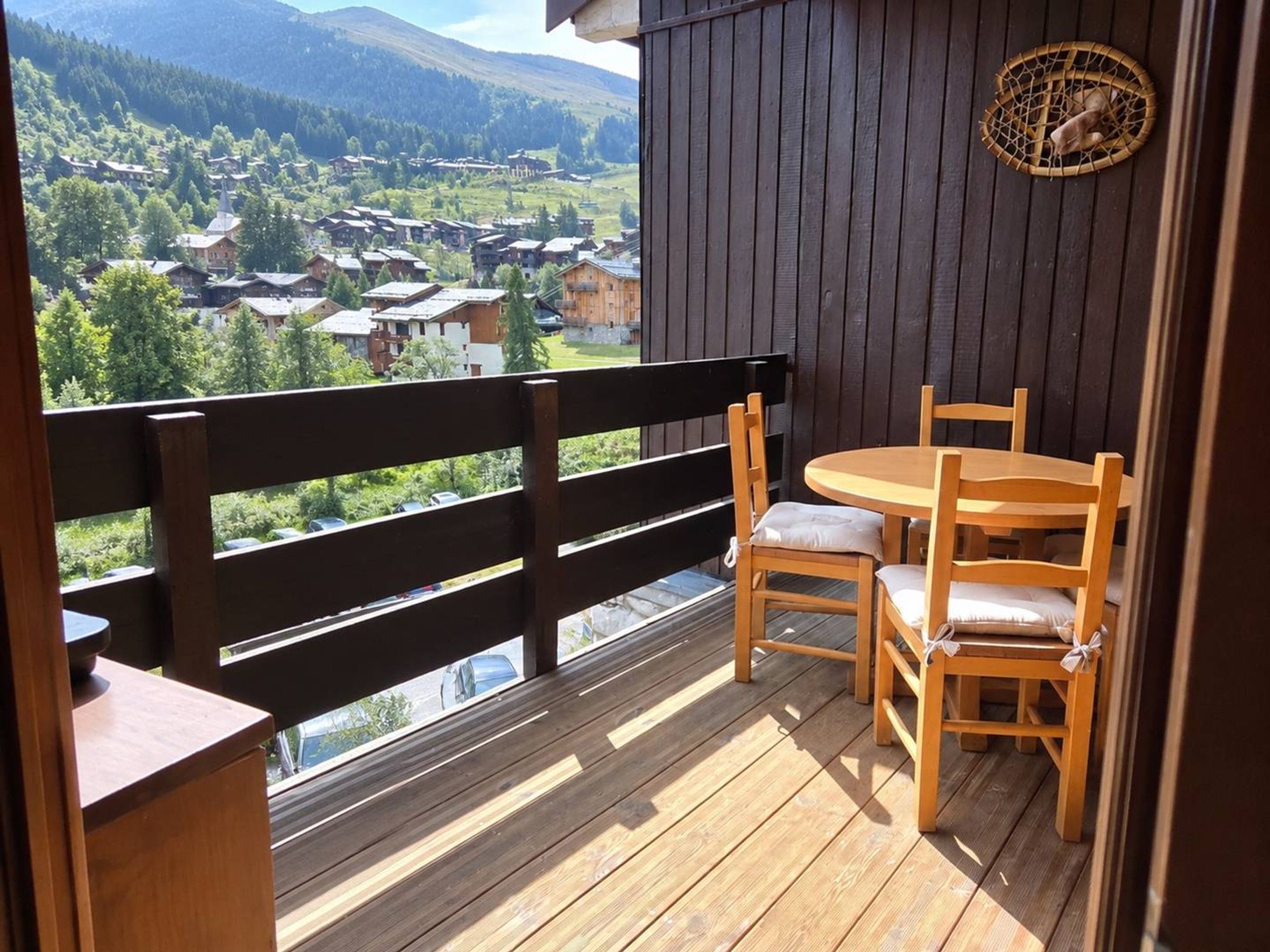 A wooden deck with a table and chairs, overlooking a village with houses and mountains in the background.