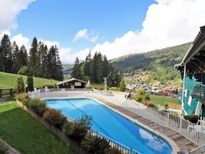 Ein Schwimmbecken mit Holzboden, umgeben von einem weißen Zaun, vor einer malerischen Landschaft aus einem Dorf und bewaldeten Hügeln.