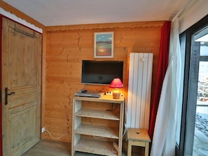Ein Holzinnenraum mit Fernseher auf einem Regal, einem kleinen Tisch mit Lampe und einem Fenster mit Vorhängen.