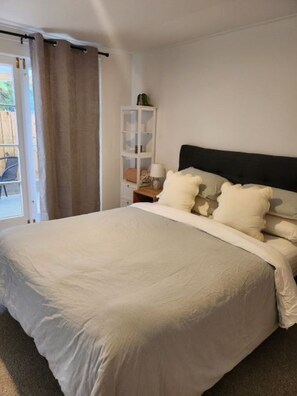 Un dormitorio con una cama grande, una mesita de noche, un estante blanco y vistas a través de una puerta.