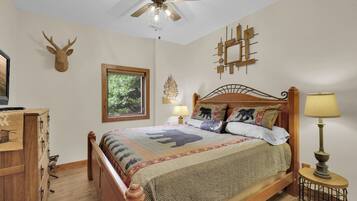 A bedroom with a wooden bed, a ceiling fan, a window with a view of greenery, a wall-mounted clock, and a bedside table with a lamp.