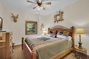 A bedroom with a wooden bed, a ceiling fan, a window with a view of greenery, a wall-mounted clock, and a bedside table with a lamp. - Cabin w Views / Hot Tub / Firepit  - close to Boone! (Vilas)