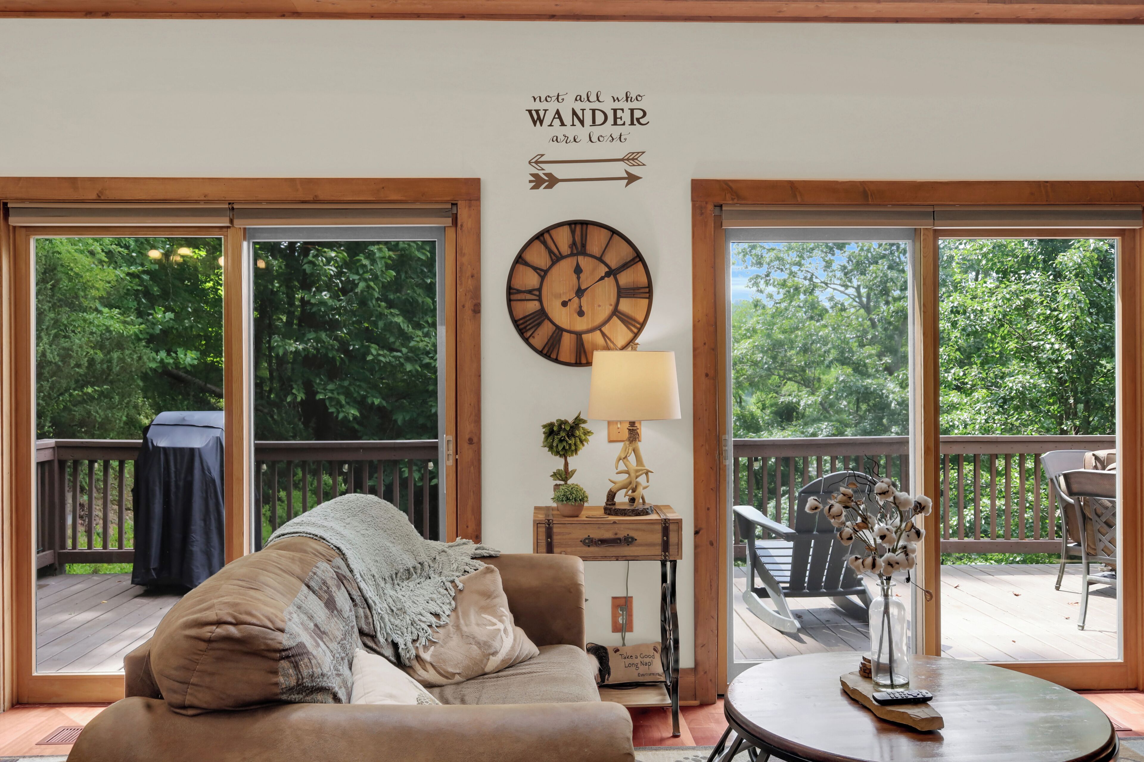 A cozy living room with a large wooden clock, a comfortable couch, and a view of a wooden deck through large glass doors.