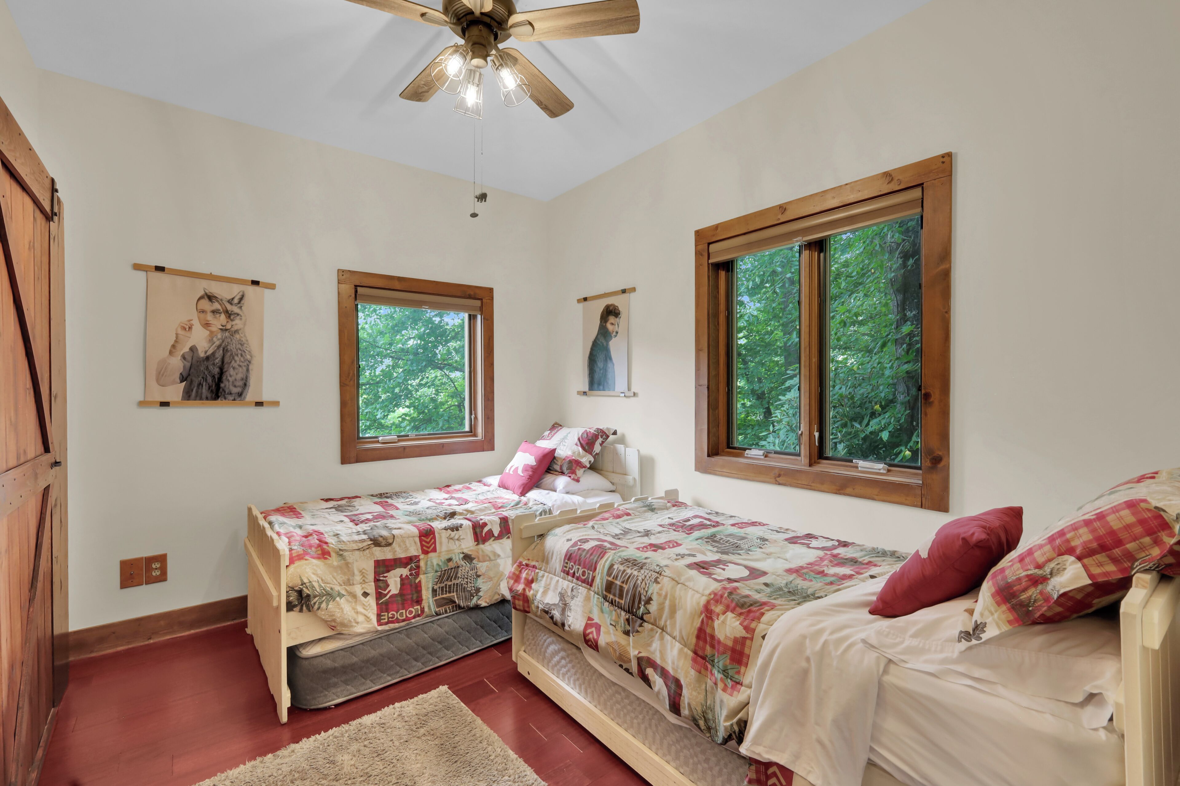 A bedroom with two beds, a ceiling fan, and two windows with views of greenery.