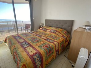 Een slaapkamer met een groot bed, een stoel bij een schuifpui, een nachtkastje met een lamp en een verwarming.