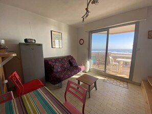 A room with a sofa, a dining table, a small TV on a cabinet, a chair, and a view of the beach through a large sliding door. - Apartment with sea view terrace - 4 guests (Canet-Plage)