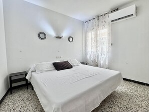Ein Schlafzimmer mit einem Bett, einem Nachttisch, einer Wanduhr, einer Wand-Klimaanlage und einem Fenster mit durchsichtigen Vorhängen.