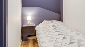 Ein kleiner, schmaler Raum mit einem Bett, einem Nachttisch und einer Lampe.