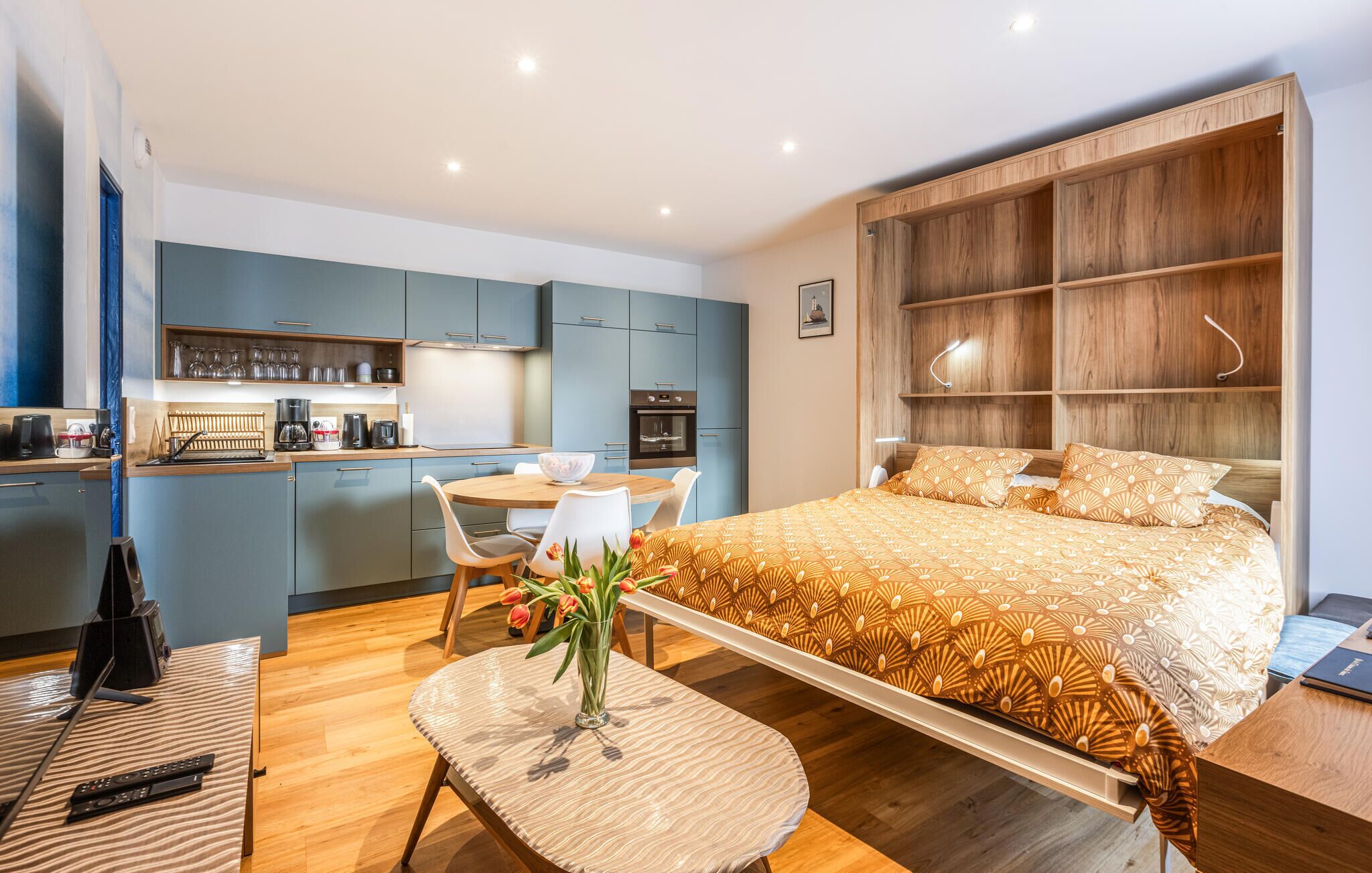 A modern hotel room with a bed, a dining table, a kitchen area, and a small balcony.