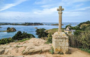 A coastal landscape with a stone cross, rocky terrain, and a view of the sea and distant buildings. - Apartment in Lezardrieux (891) (Lezardrieux)