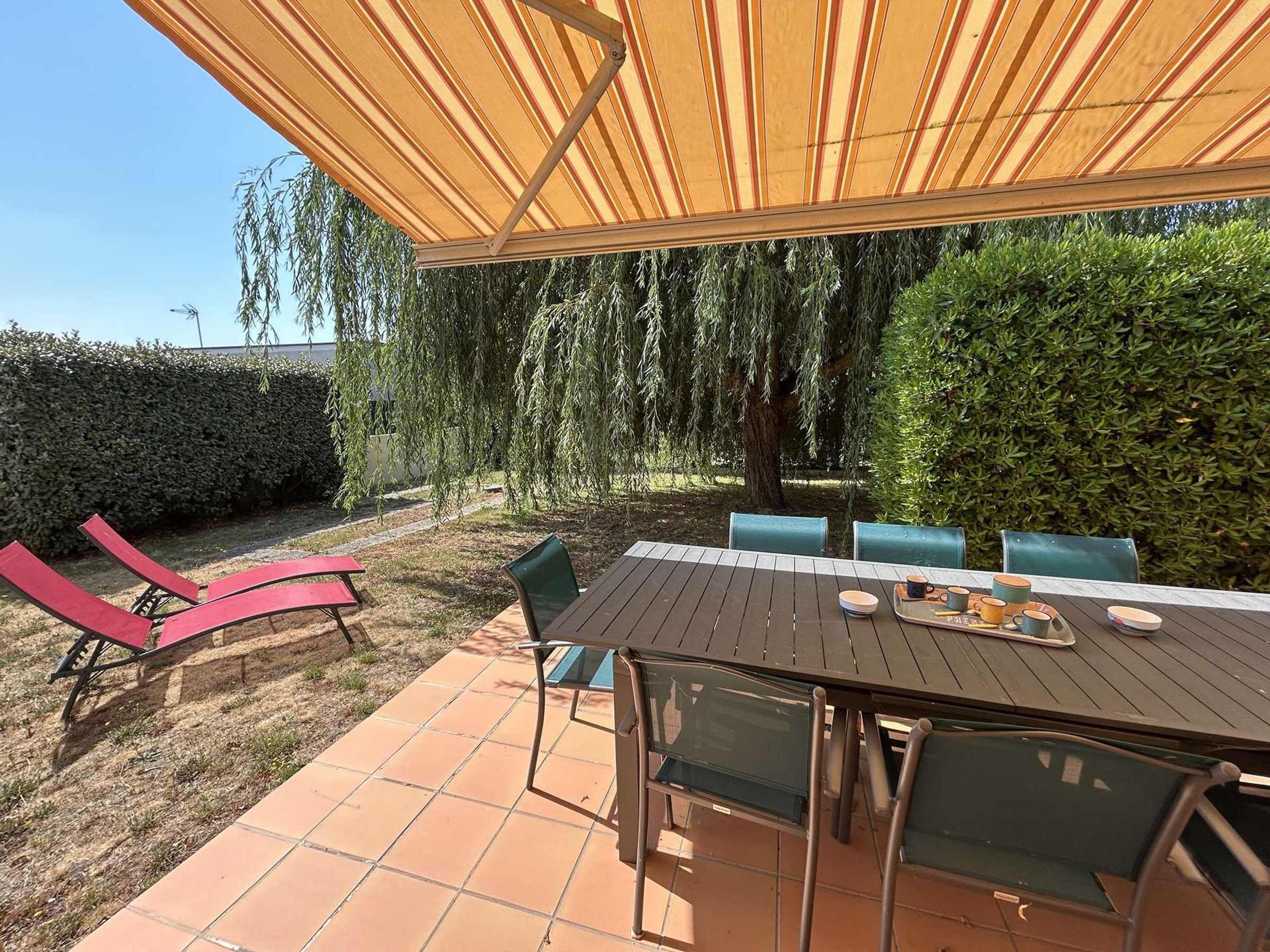 Un patio al aire libre con una mesa y sillas, un toldo a rayas y un seto de fondo.
