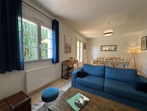 Ein Wohnzimmer mit einem blauen Sofa, einem Holztisch mit einem Buch, einem kleinen Hocker und einem Fenster mit Vorhängen.