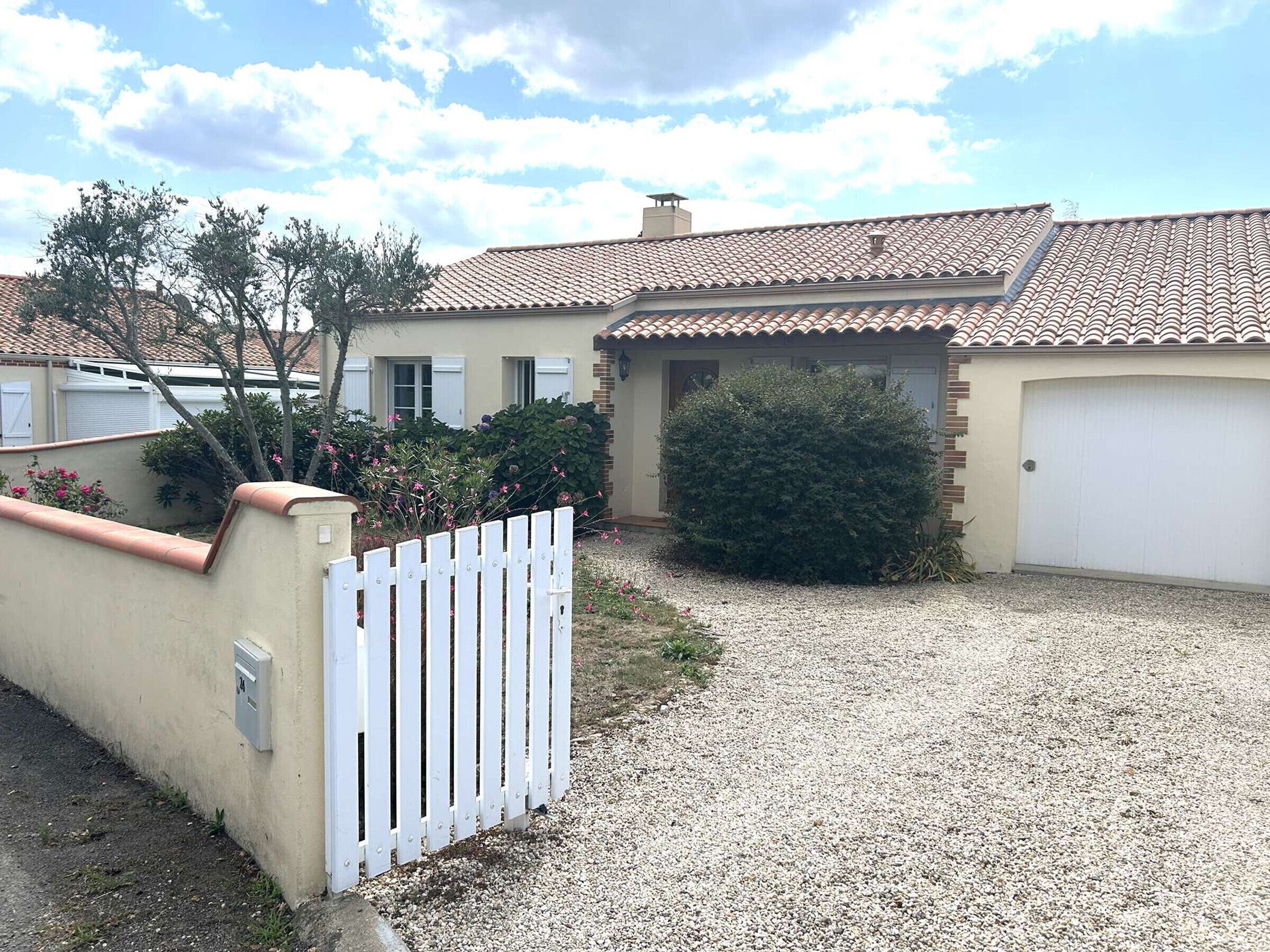 Una propiedad residencial con camino de entrada de grava, cerca de piquetes blancos y un pequeño jardín con arbustos y flores.