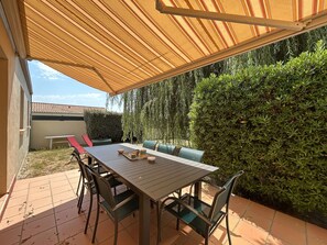 Eine Terrasse im Freien mit Tisch und Stühlen, einem überdachten Bereich und einer Hecke.