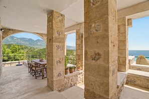 Ein von Steinbögen geschützter Außenbereich mit Tisch und Stühlen, Blumentöpfen und Blick auf das Meer und die Berge.