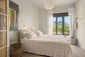 Ein Schlafzimmer mit Bett, Nachttisch, Lampe, einem Fenster mit Vorhängen und Blick ins Freie.
