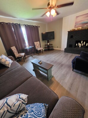 A living room with a sofa, chairs, a coffee table, a flat-screen TV, a fireplace, and a ceiling fan. - Charming 3 bedroom house in St Robert. Only 3 minutes from the main gate of FLW. (Saint Robert)
