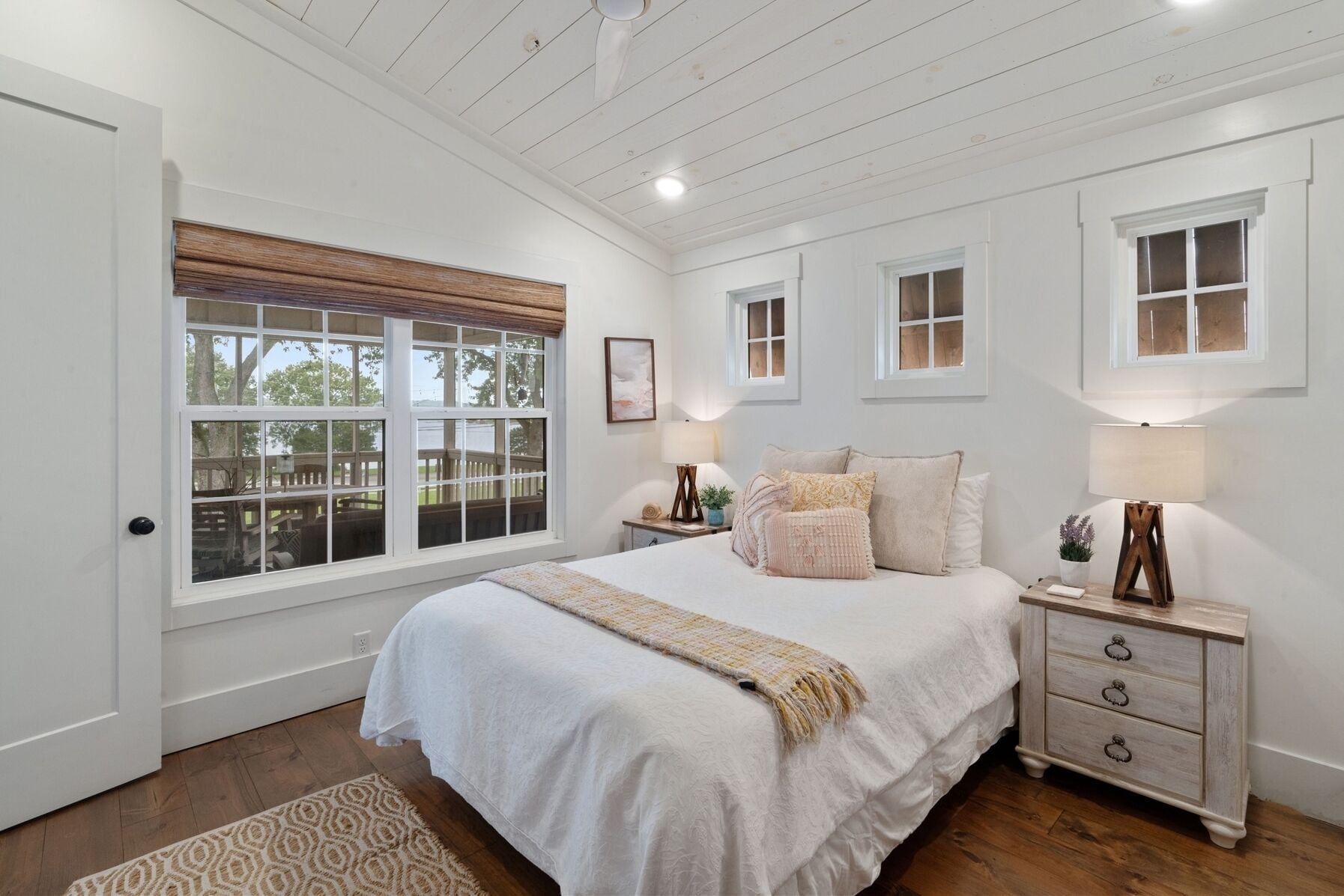 A bedroom with a large bed, two bedside tables, a window with blinds, and a wooden ceiling.